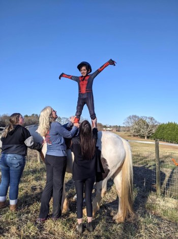 Instructors help a participant stand on one of the horses at the farm.